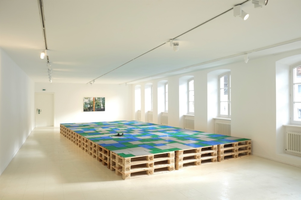 Ausstellungsansicht Natascha Sadr Haghighian "le lion malade" Blick in einen Ausstellungsraum. Mittig steht eie Installation aus Paletten, mit aufgesetzten Legoplatten, Form und Größe erinnert an einen Panzer. Hinten eine großformatige Fotografie.