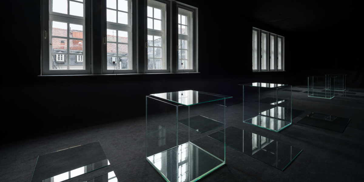 Matej Bosnić – πάσχειν (páskhein), 2021, Detail Ausstellung Stadtgalerie Blick in einen schwarzen Raum. Durch die Fenster scheint Licht. Auf dem Boden stehen vier Glaskuben