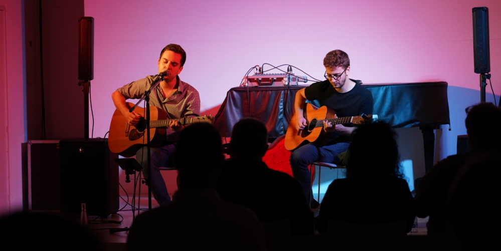 Stadtgalerie Draußen Konzert von Lukas Schüßler und Phillipp Herget Veranstaltungsraum der Stadtgalerie, Umrisse von einem Publikum, zwei männliche Gitarrenspieler auf der Bühne