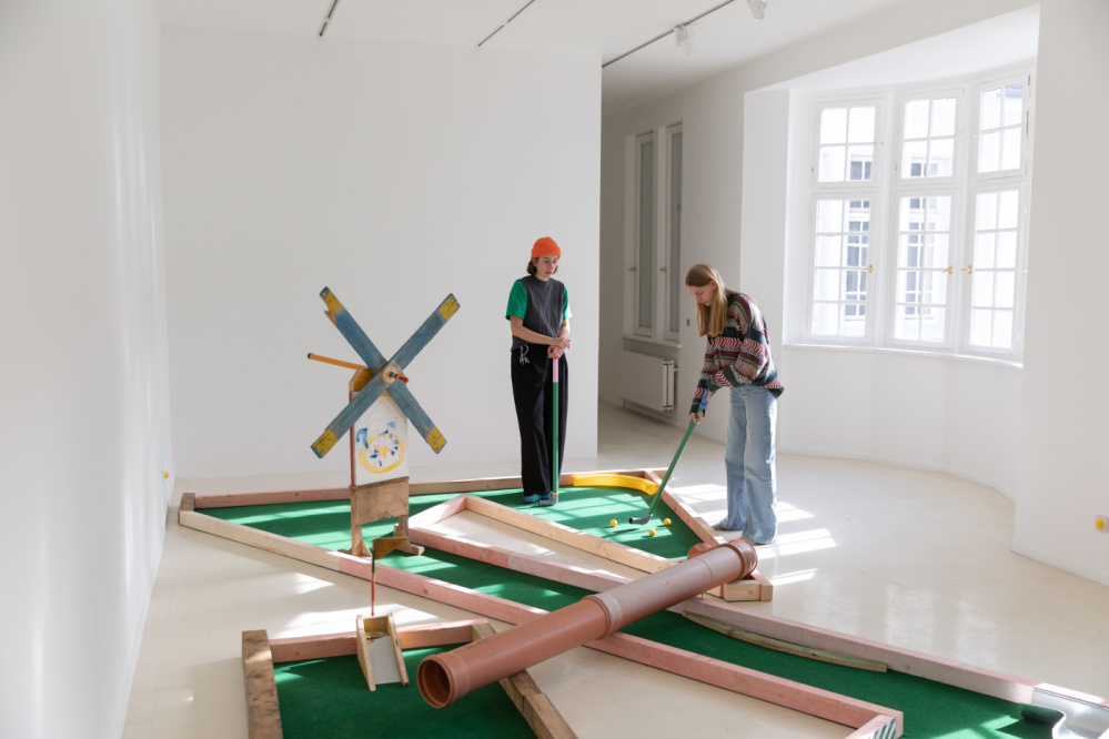 Driving Range - Jonas Mayer Eine selbstgebaute Minigolfbahn. Gebaut aus Holzbalken und Baurohren. Zwei Personen, die mit Schlägern aus Kunststoffrohren Minigolf spielen.