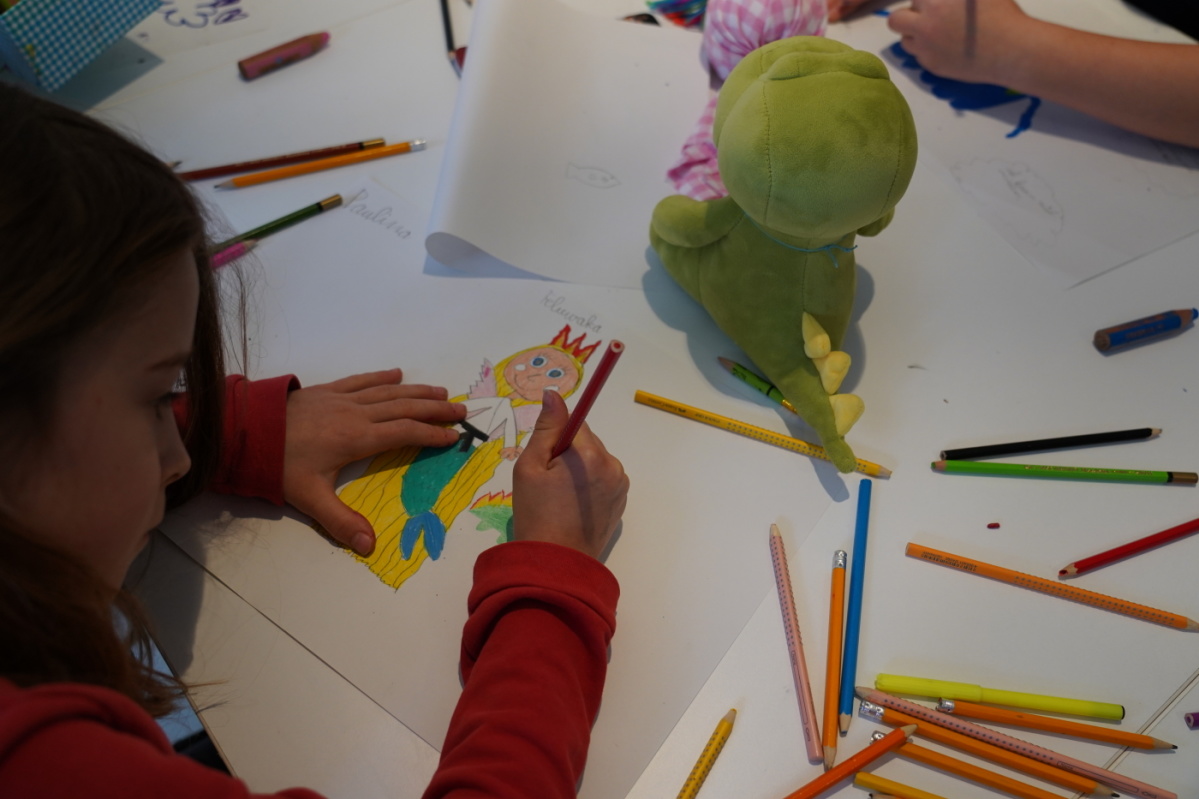Kindersonntag mit Hannah Mevis und Noé Duboutay Buntstifte und buntbemalte Blätter auf einem Tisch. In der Mitte ein grünes Kuscheltier in Form eines Dinos