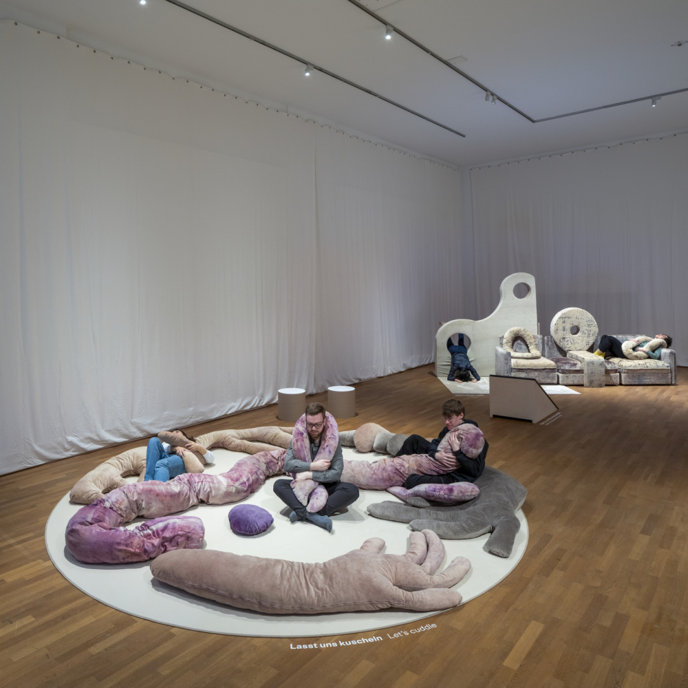 Hamburger Museum für Kunst und Gewerbe, Ausstellungsansicht, Well Beings, Valentina Karga, 2023 Ausstellungsraum mit Installationen, Plüsch Tiere, Kissen, Sofa und Besucher*innen