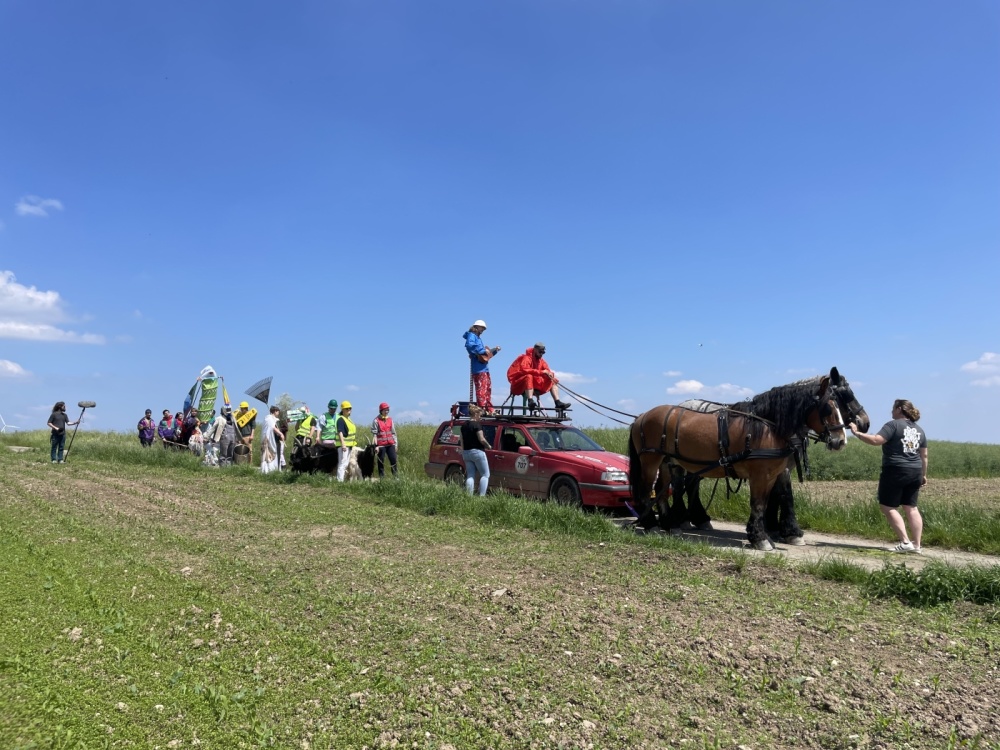 Experimental- und Kunstprojekt La Capsule, Mai - August 2024 Auf einer Landstraße Parade mit verkleideten Personen, zwei Pferde, die einen Wagen ziehen