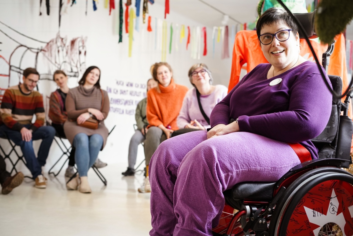 Inklusionsprojekt Frau in Rollstuhl sitzt vor einer Gruppe von Menschen, alle schauen begeistert in Richtung der Kamera aber an der Kamera vorbei