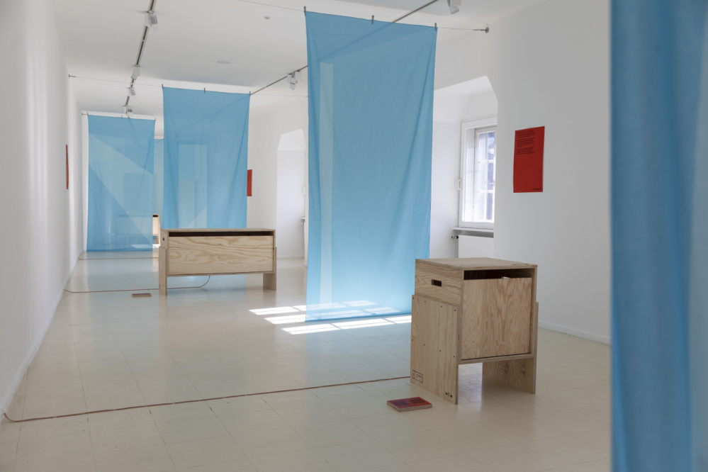 TRANSIT Mark Braun Ausstellungsansicht TRANSIT von Mark Braun, zwei Sperrholzkisten und leichte blaue Vorhänge im weißen Galerieraum