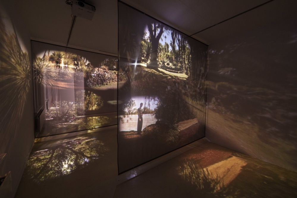 LOOPZONES Suzan Noesen Blick in einen abgedunkelten Ausstellungsraum in dem auf zwei transparenten Leinwänden ein Film zu sehen ist